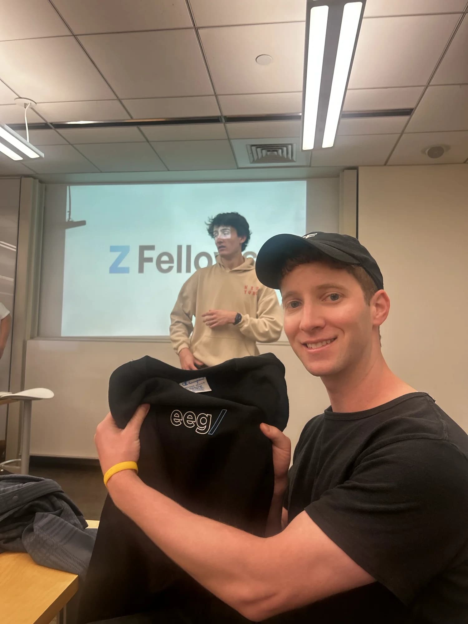 A member holding up an EEG crewneck in a classroom, a talk going on behind