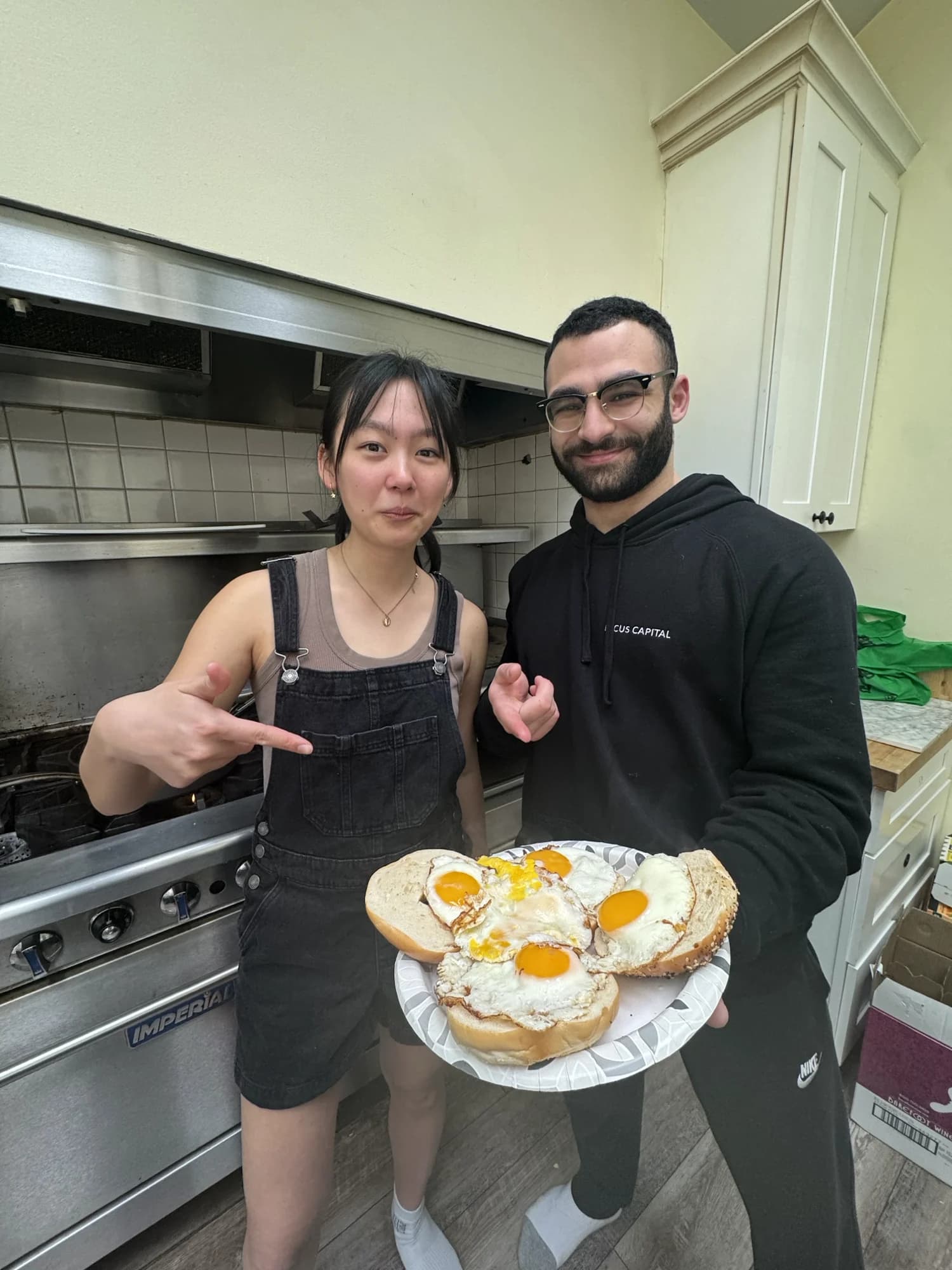 Two members presenting a plate of fried-egg sandwiches in a kitchen