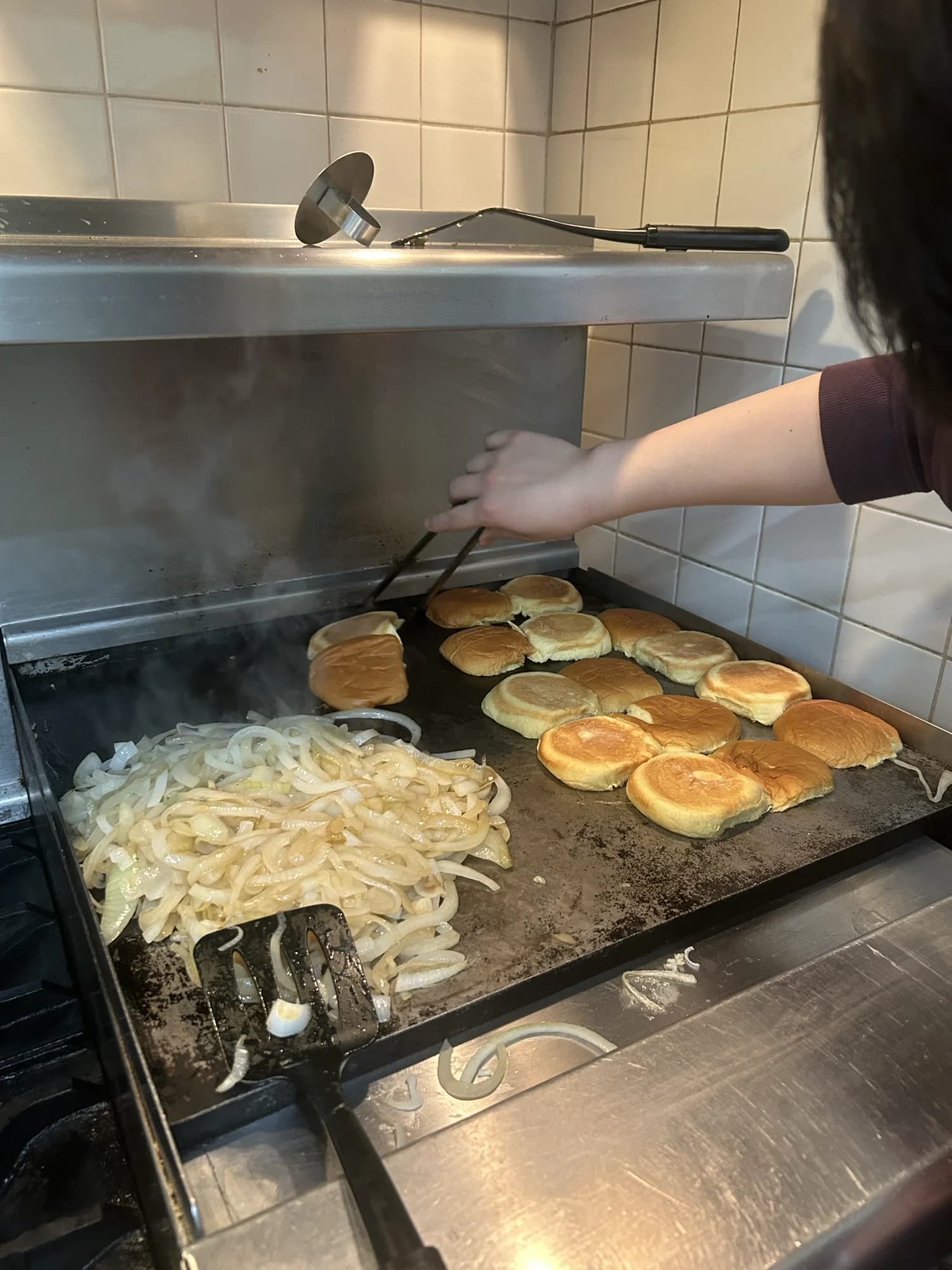 Buns and onions toasting on a flat-top griddle, a hand working the tongs