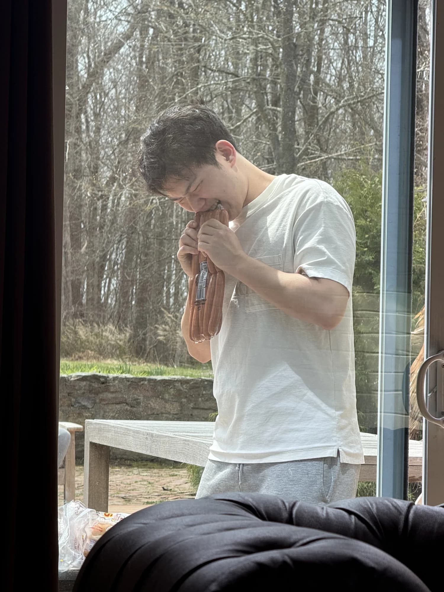 A member eating a link of sausage by a window looking onto bare woods