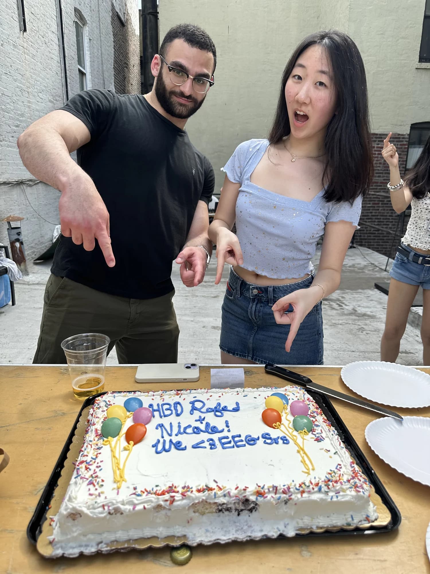 Two members pointing at a sheet cake reading HBD Rajan and Nicole, we love EEG srs